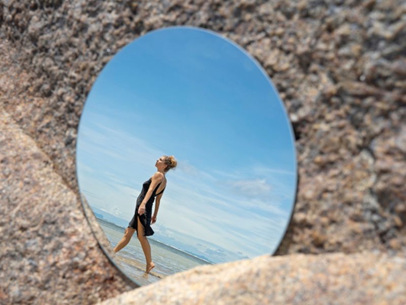woman on beach viewed on circular mirror resting on rocks